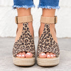 A pair of women's wedge sandals with a beige and brown leopard print upper, ankle straps with buckles, and braided jute platform soles, shown on a model's feet with frayed blue jeans.