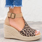 A close-up of a person's foot wearing a wedge espadrille sandal with a leopard print upper and a beige ankle strap with a metal buckle, paired with frayed blue jeans.