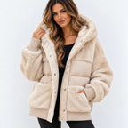 A woman wearing a beige sherpa-lined puffer jacket with a plush hood, ribbed cuffs, and front pockets, styled over a black top against a light gray background.