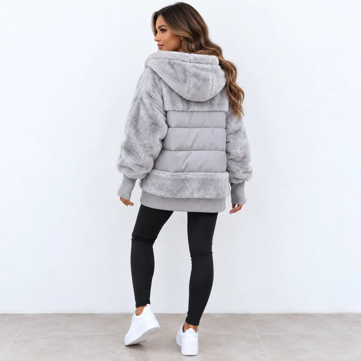 A woman wearing a light gray fuzzy hooded puffer jacket, black leggings, and white sneakers, shown from the back against a plain white wall.