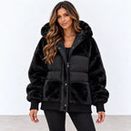 A woman wearing a black faux fur puffer jacket with a large plush hood, ribbed cuffs and hem, and silver snap buttons, standing against a plain white studio background.