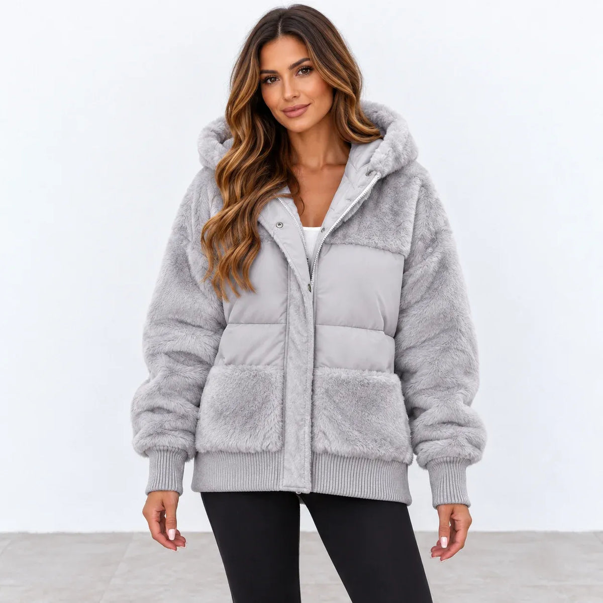 A woman modeling a light gray hooded puffer jacket with a faux-fur texture, ribbed cuffs and hem, and a front zipper, paired with black pants against a plain white studio background.