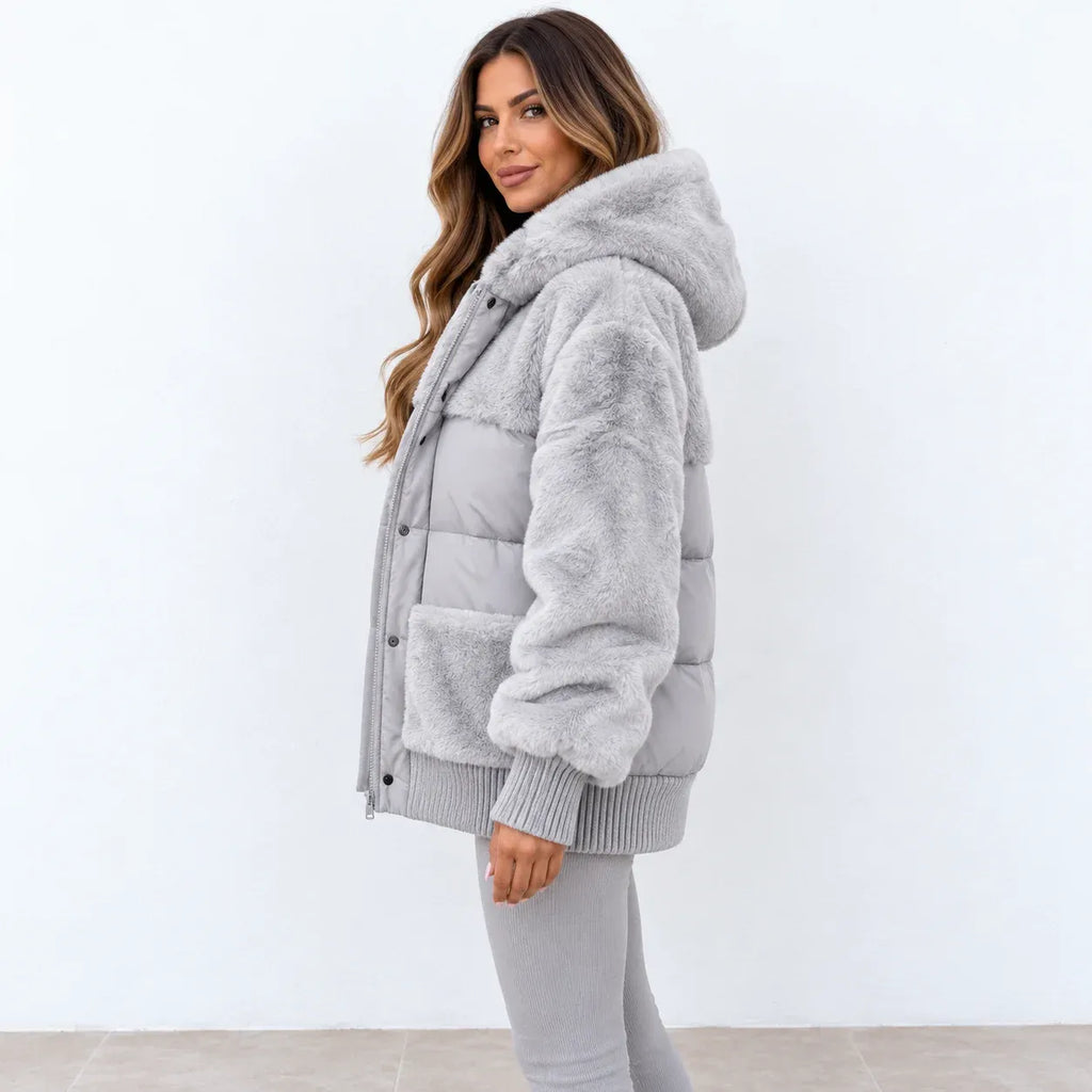 A woman wearing a light gray faux fur puffer jacket with a hood and ribbed knit trim, paired with gray leggings, standing against a plain white wall.