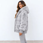 A woman wearing a light gray faux fur puffer jacket with a hood and ribbed knit trim, paired with gray leggings, standing against a plain white wall.
