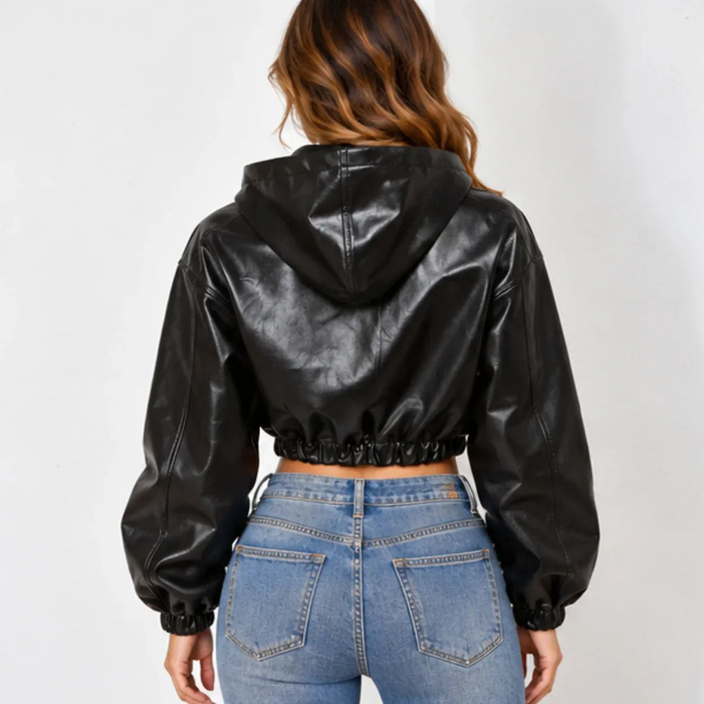 A woman viewed from the back wearing a black faux-leather cropped hooded jacket with long sleeves and elastic cuffs, paired with blue jeans, against a plain light gray background.