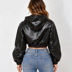 A woman viewed from the back wearing a black faux-leather cropped hooded jacket with long sleeves and elastic cuffs, paired with blue jeans, against a plain light gray background.