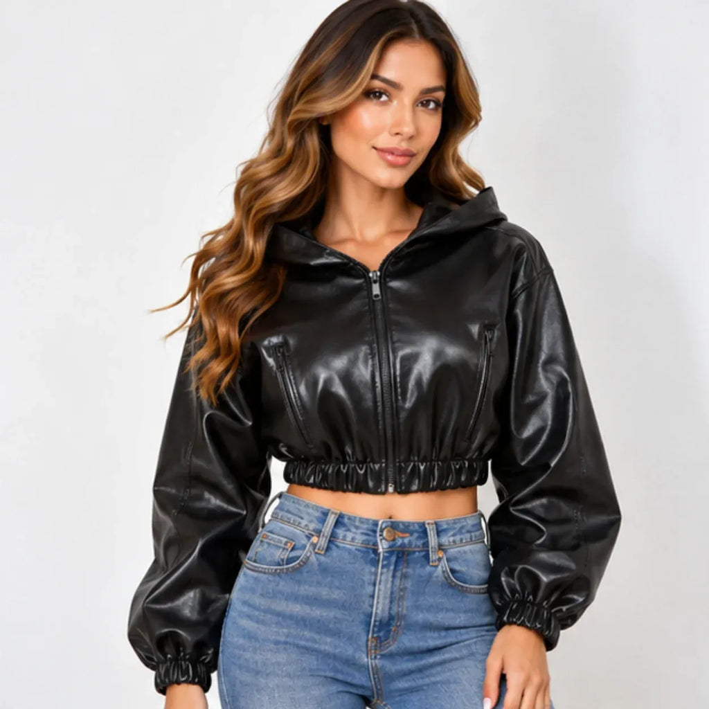 A woman modeling a black faux-leather cropped zip-up hoodie with long sleeves and elastic cuffs, paired with blue jeans, against a light gray studio background.