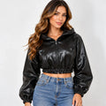 A woman modeling a black faux-leather cropped zip-up hoodie with long sleeves and elastic cuffs, paired with blue jeans, against a light gray studio background.