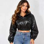 A woman modeling a black faux-leather cropped zip-up hoodie with long sleeves and elastic cuffs, paired with blue jeans, against a light gray studio background.