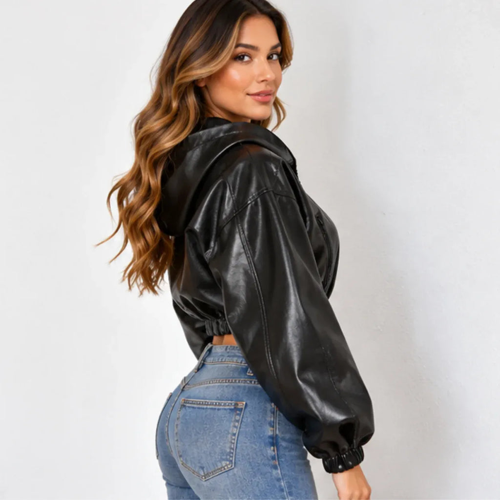 A woman wearing a black faux-leather cropped hooded jacket with balloon sleeves and elastic cuffs, paired with blue jeans, photographed against a plain light gray wall.
