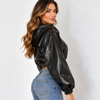 A woman wearing a black faux-leather cropped hooded jacket with balloon sleeves and elastic cuffs, paired with blue jeans, photographed against a plain light gray wall.