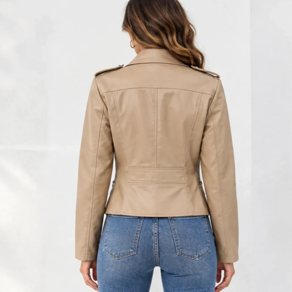 A woman viewed from the back wearing a cropped tan faux-leather jacket with shoulder epaulets and blue jeans, set against a plain light gray studio background.