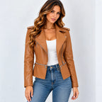 A woman modeling a cropped tan faux-leather moto jacket with silver zippers, worn over a white tank top and blue jeans against a light gray background.