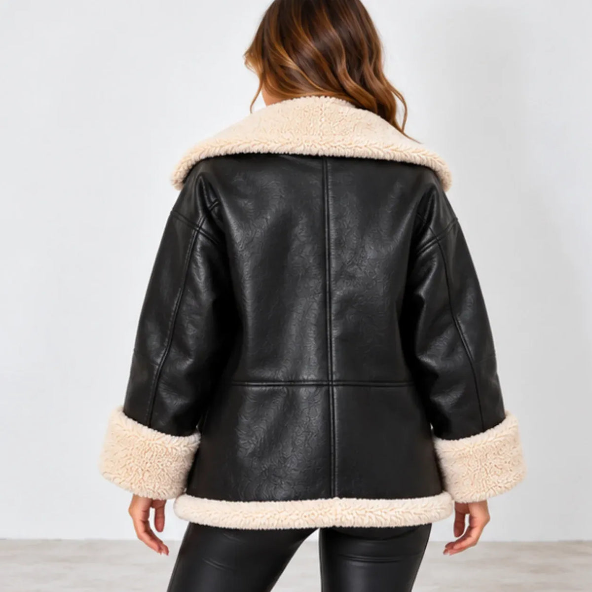 A woman shown from the back wearing a black faux-leather shearling jacket with a cream-colored plush collar, cuffs, and hem, paired with black leggings against a light gray studio background.