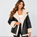 A woman modeling a black faux-leather shearling jacket with a thick cream-colored fleece lining. She is wearing a white crop top and black pants against a plain light gray studio background.