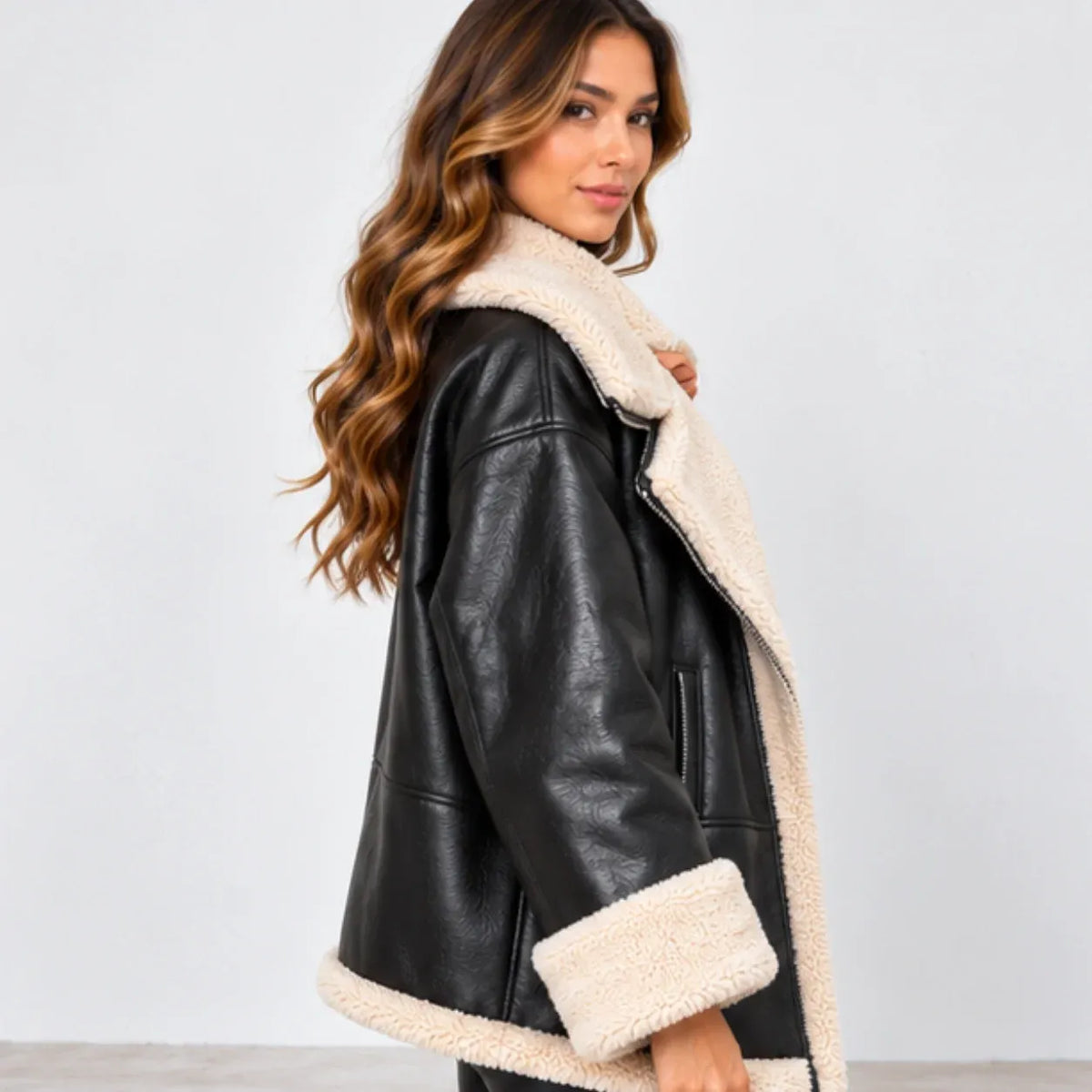 A woman wearing a black faux-leather shearling jacket with a thick cream-colored sherpa lining, photographed against a plain light gray studio background.