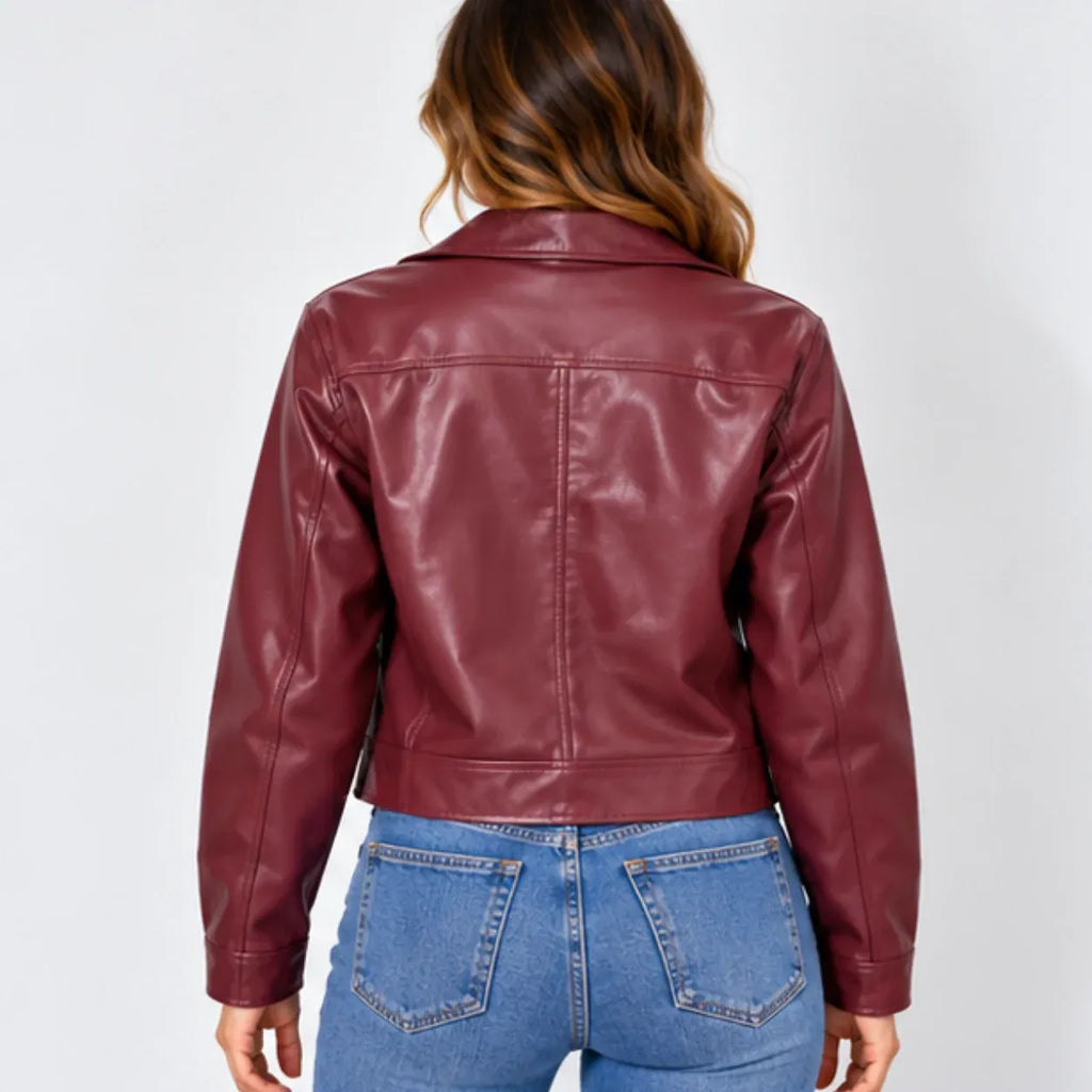 A woman viewed from the back wearing a cropped, glossy burgundy faux-leather jacket and blue jeans against a light gray studio background.