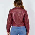 A woman viewed from the back wearing a cropped, glossy burgundy faux-leather jacket and blue jeans against a light gray studio background.