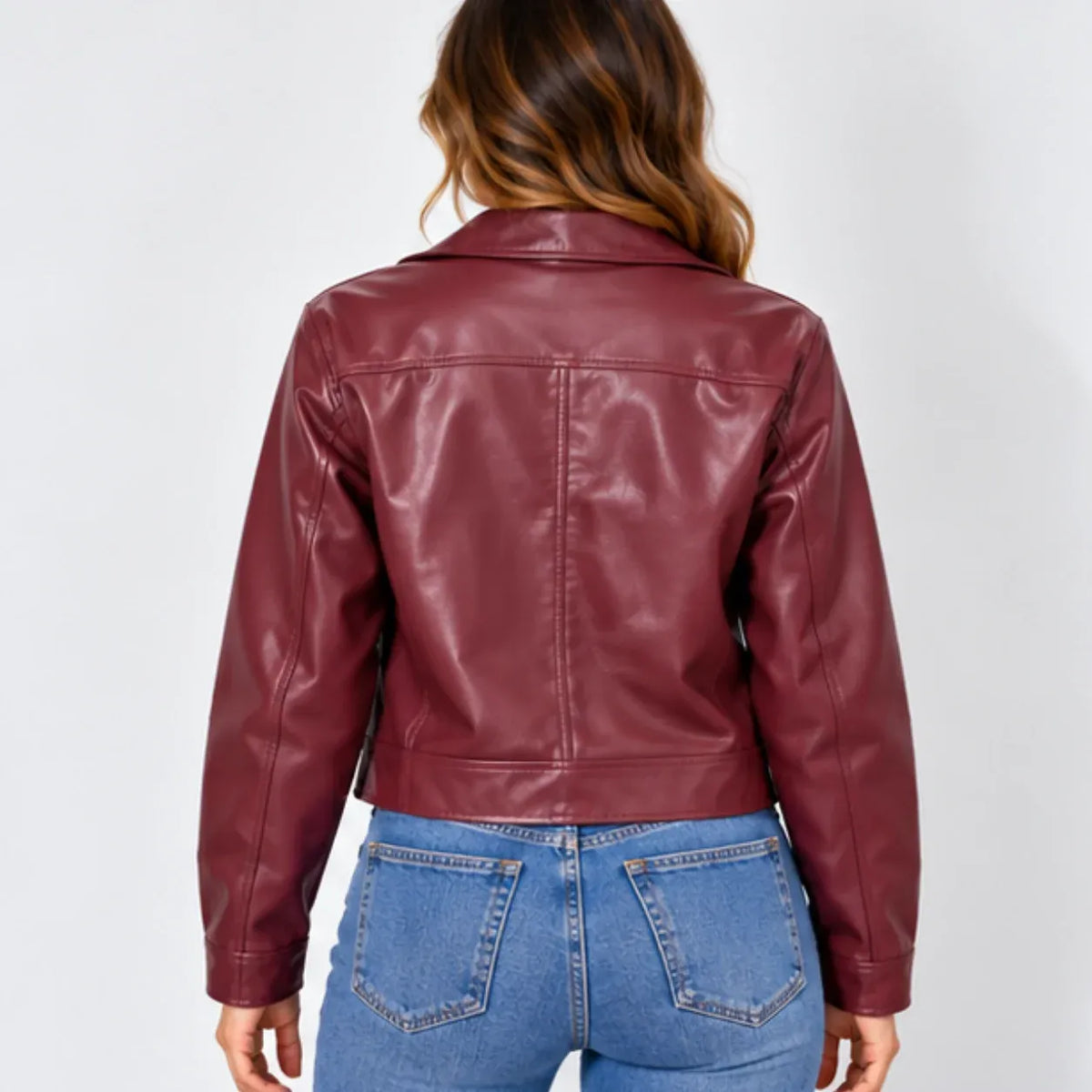 A woman viewed from the back wearing a cropped, glossy burgundy faux-leather jacket and blue jeans against a light gray studio background.