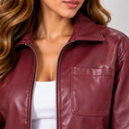 A close-up of a woman wearing a burgundy faux leather jacket with a collar, front zipper, and a chest pocket over a white ribbed top.