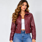 A woman modeling a cropped, long-sleeve faux leather jacket in a deep burgundy color, worn over a white crop top and blue jeans against a light gray studio background.