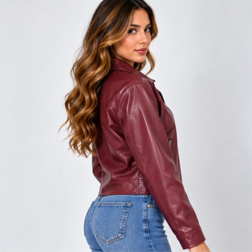 A woman with long wavy brown hair is shown from the side and back, wearing a cropped, glossy burgundy faux-leather jacket and blue jeans against a light gray studio background.