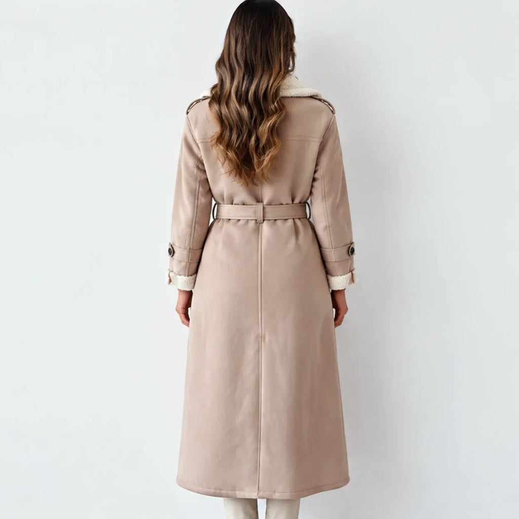A woman viewed from behind wearing a long beige trench coat with a cream collar and cuffs, photographed against a plain light gray studio background.