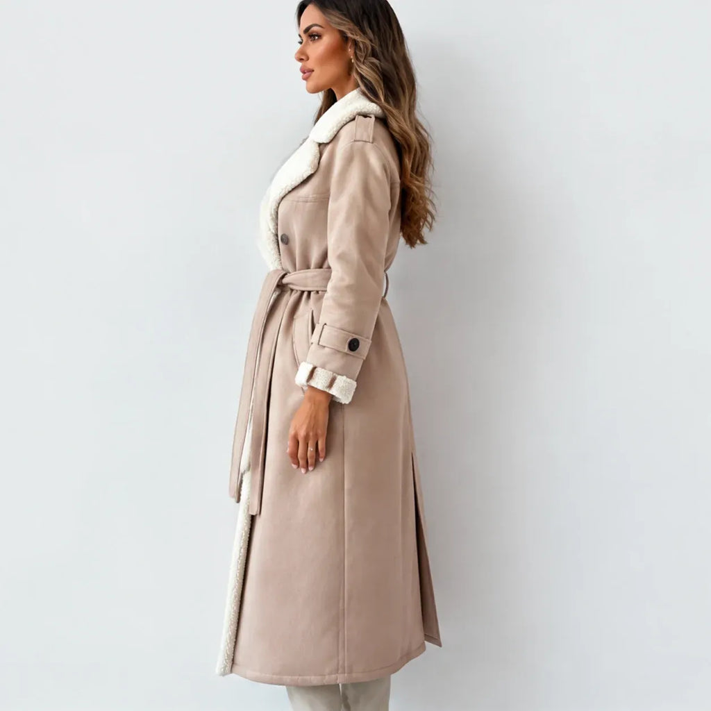 A woman wearing a long beige trench coat with a white faux-fur collar and lining, standing in profile against a light gray studio background.