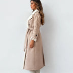 A woman wearing a long beige trench coat with a white faux-fur collar and lining, standing in profile against a light gray studio background.