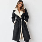 A woman modeling a black faux-suede trench coat with a thick cream sherpa lining. The coat features a matching waist tie belt, wide lapels, and a plush collar and cuffs, shown against a plain light gray background.