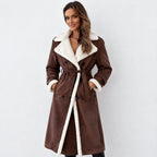 A woman modeling a brown faux-suede shearling coat with a white sherpa lining, featuring a double-breasted design, belted waist, and collar, set against a plain light gray background.