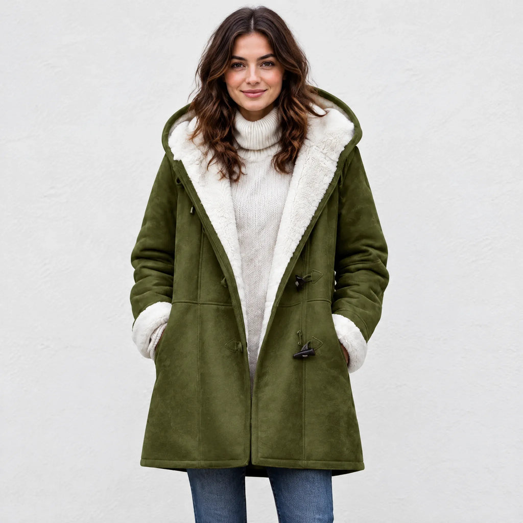 A woman wearing an olive green hooded shearling coat over a cream turtleneck sweater and blue jeans, standing against a light gray textured wall.