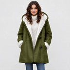 A woman wearing an olive green hooded shearling coat over a cream turtleneck sweater and blue jeans, standing against a light gray textured wall.