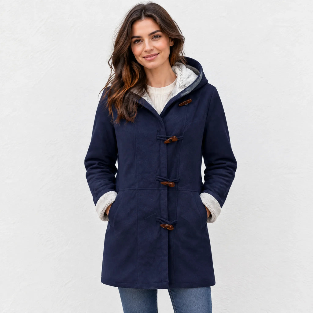 A woman wearing a navy blue hooded duffle coat with a light gray sherpa lining and brown toggle closures, standing against a white textured wall.