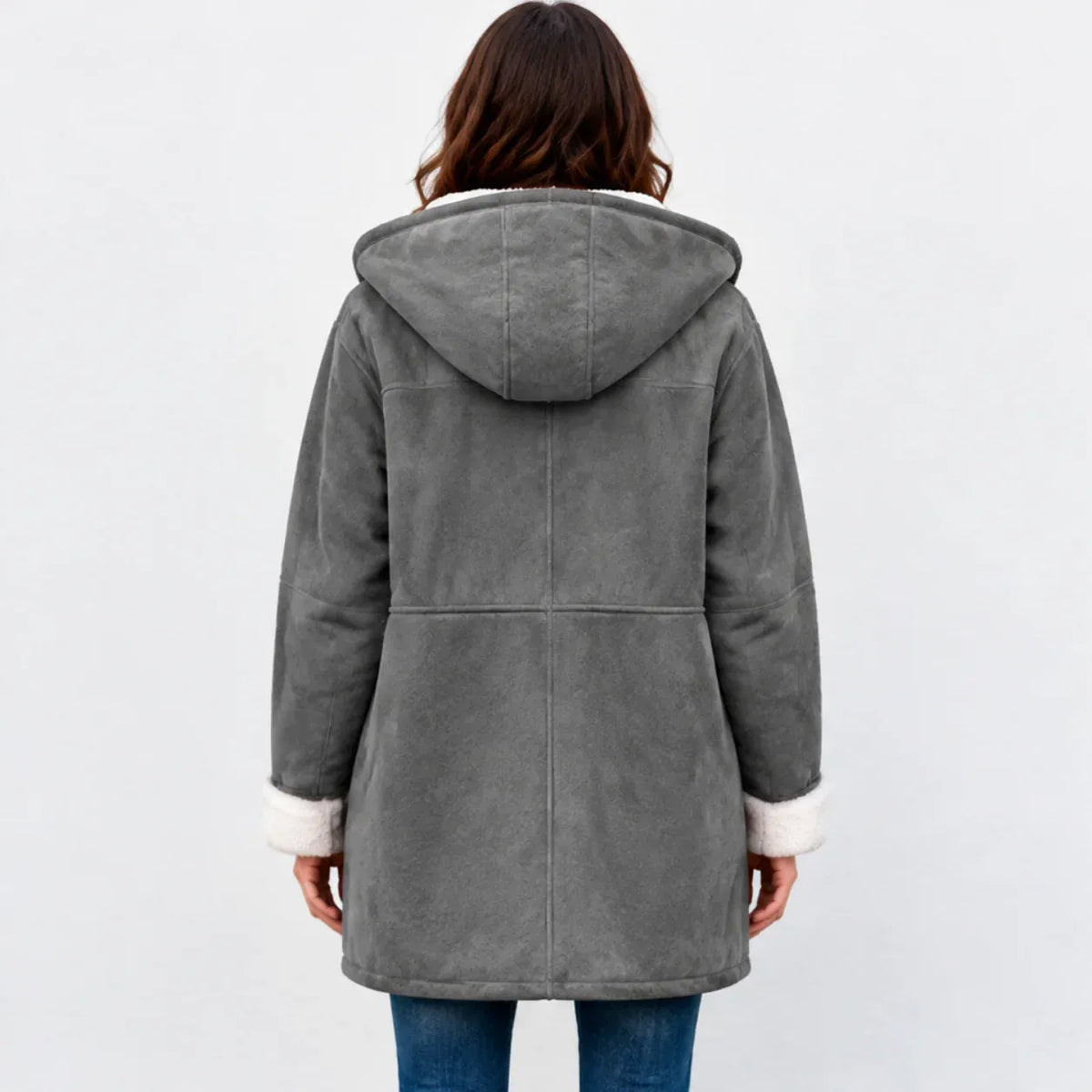 A woman viewed from the back wearing a gray hooded shearling-style coat with white faux-fur trim at the hood and cuffs, paired with blue jeans against a light gray studio background.