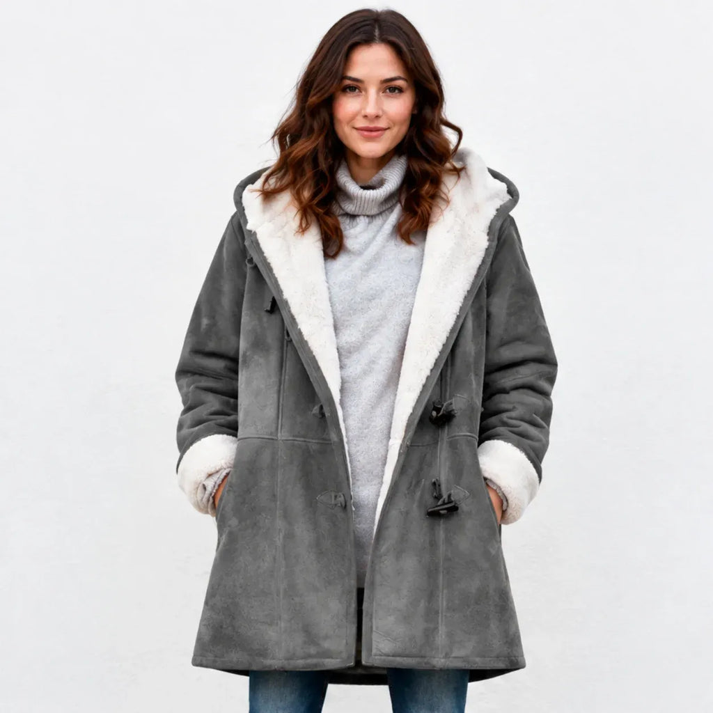 A woman wearing a gray suede-like hooded coat with a white sherpa lining, styled over a light gray turtleneck sweater and blue jeans, against a plain white studio background.