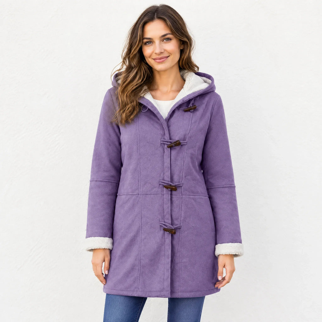 A woman wearing a purple hooded duffle coat with a white sherpa lining, paired with blue jeans, standing against a white textured wall.
