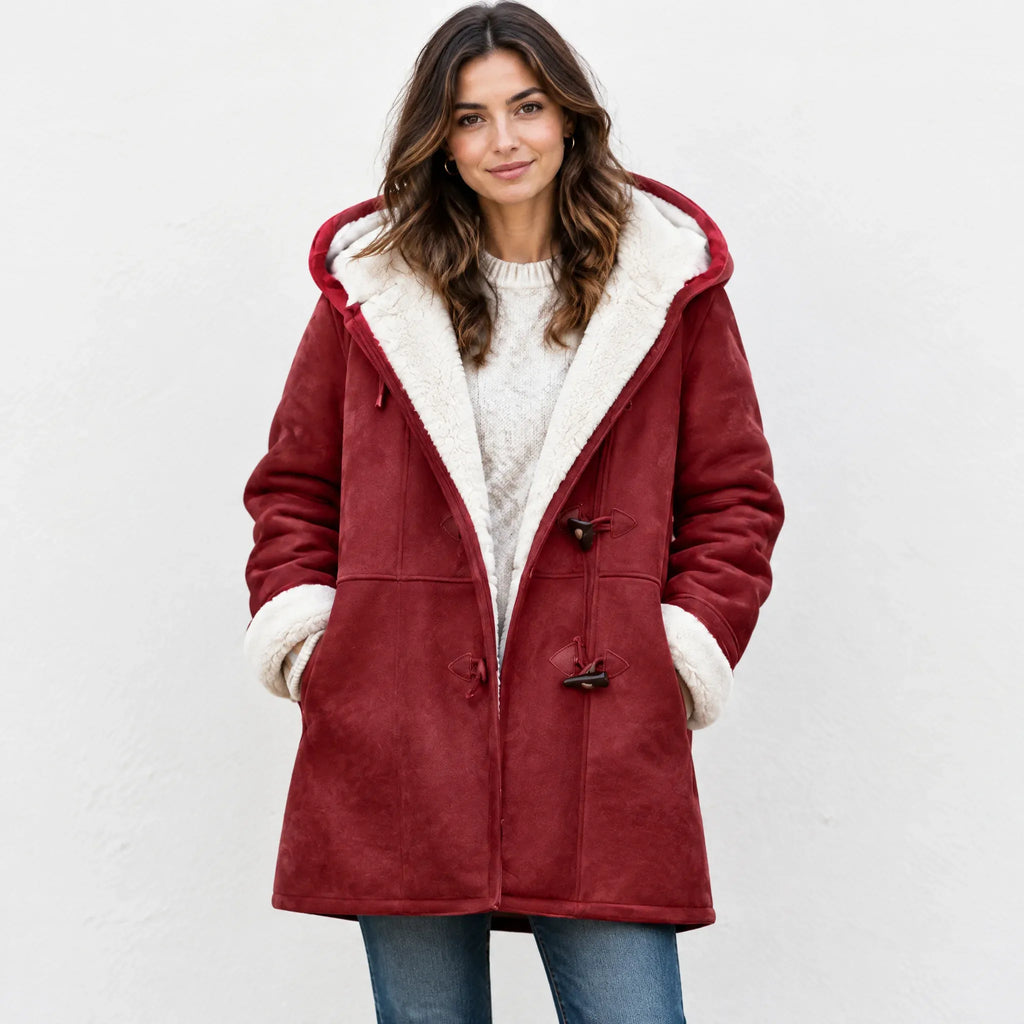 A woman wearing a burgundy faux-suede hooded coat with a cream sherpa lining, paired with a cream knit sweater and blue jeans, standing against a light gray wall.
