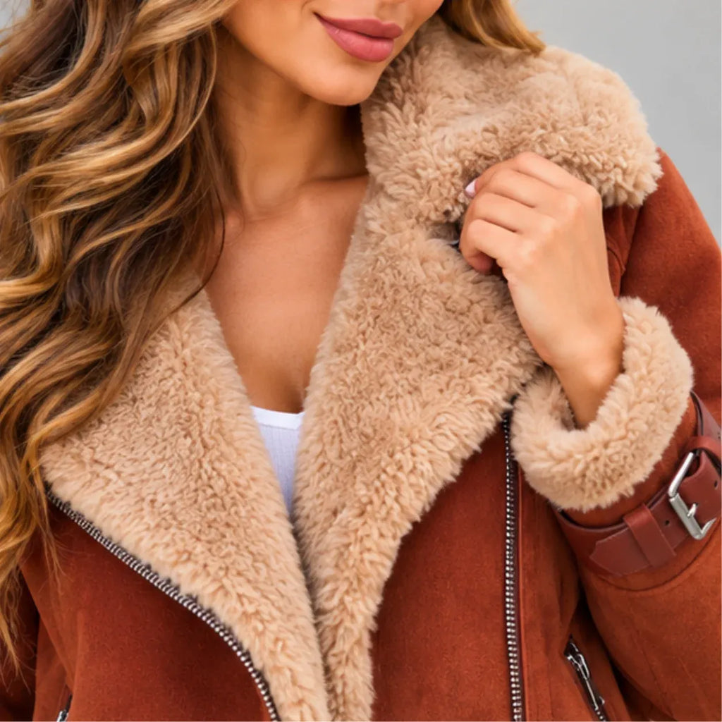 A close-up of a woman wearing a brown suede-like jacket with a thick beige sherpa fleece collar and cuffs, holding the collar open. The jacket has silver zippers and a brown belt detail on the sleeve.