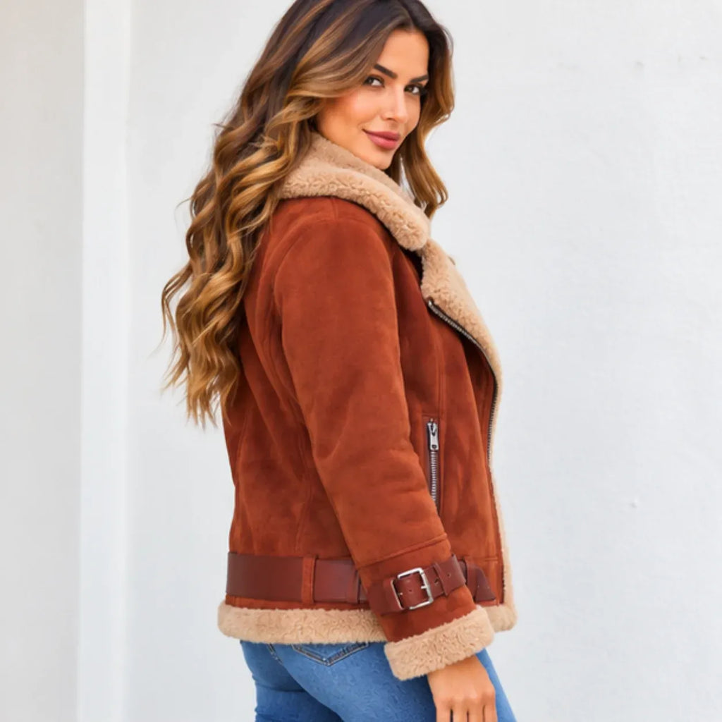 A woman wearing a brown suede shearling jacket with a tan sherpa lining, paired with blue jeans, photographed against a plain white wall.