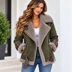 A woman wearing an olive green shearling-lined aviator jacket with brown faux-leather trim and belt details, paired with a white top and blue jeans, standing in front of a white building.