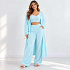 A woman models a light blue fuzzy lounge set consisting of a cropped tank top, matching wide-leg pants, and a long open-front cardigan, styled with fluffy slip-on slippers in a bright, sunlit room.