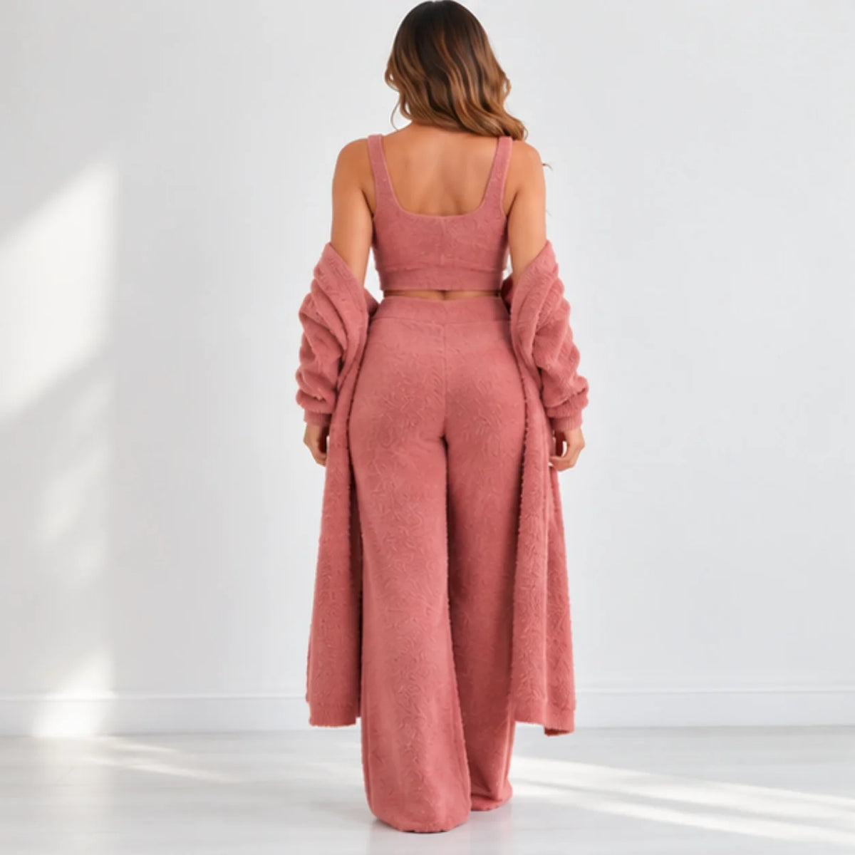 A woman stands with her back to the camera wearing a dusty rose two-piece outfit consisting of a cropped tank top and wide-leg pants, styled with a matching fuzzy open-front cardigan draped off her shoulders.