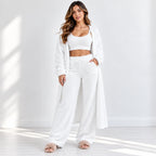 A woman modeling a cozy white lounge set consisting of a cropped tank top, matching wide-leg pants, a long open-front cardigan, and fuzzy slide sandals, set against a bright white studio background with soft window light shadows.