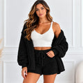 A woman modeling a cozy two-piece loungewear set consisting of a black sherpa zip-up cardigan and matching shorts, worn over a white fuzzy camisole top, set against a white paneled wall.