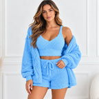 A woman modeling a light blue fuzzy three-piece loungewear set, consisting of a cropped camisole top, matching shorts with a drawstring, and an oversized zip-up cardigan, set against a white paneled wall.