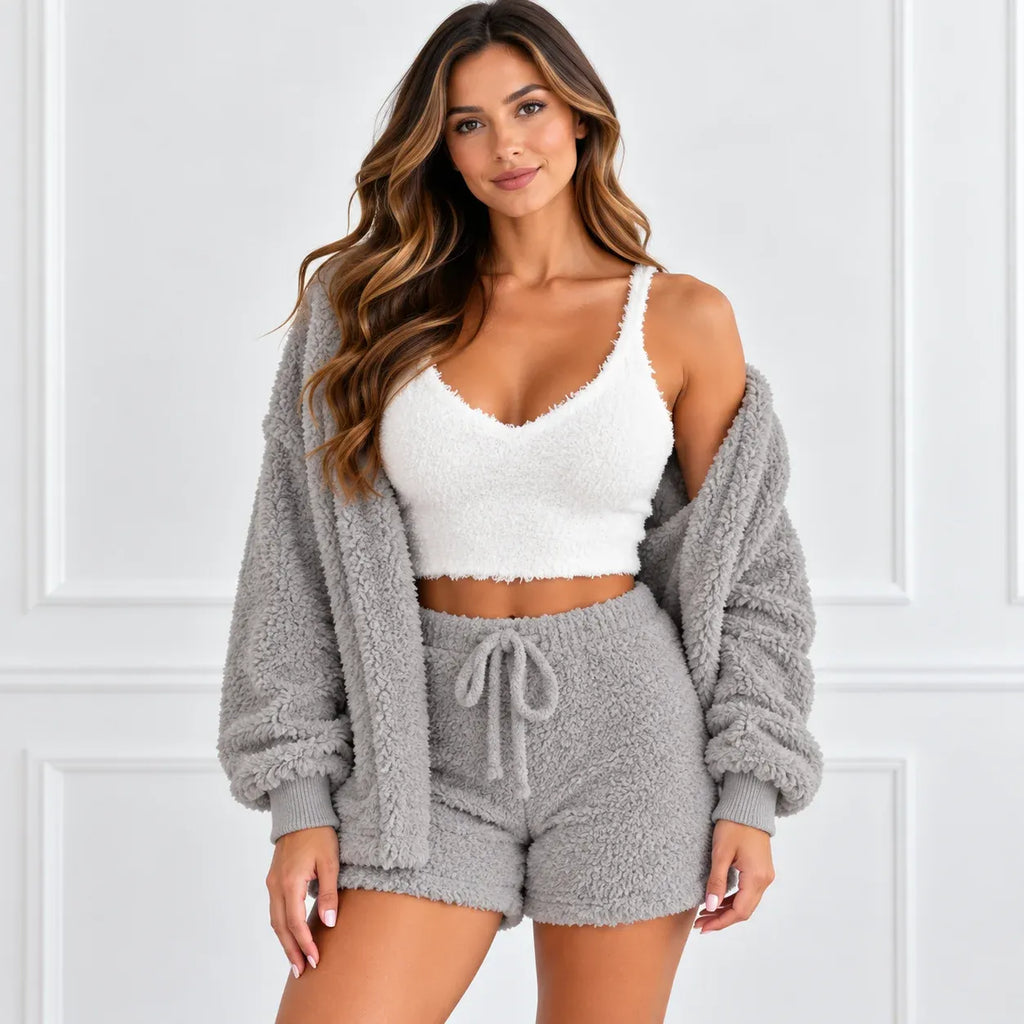 A woman modeling a cozy two-piece loungewear set consisting of a white fuzzy crop top and a matching gray fuzzy drawstring shorts, styled with an open gray fuzzy cardigan against a white paneled wall.