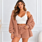 A woman models a cozy three-piece fleece loungewear set consisting of a matching cropped cardigan, bralette, and shorts in a dusty rose-brown color, paired with a white cropped camisole against a white paneled wall.