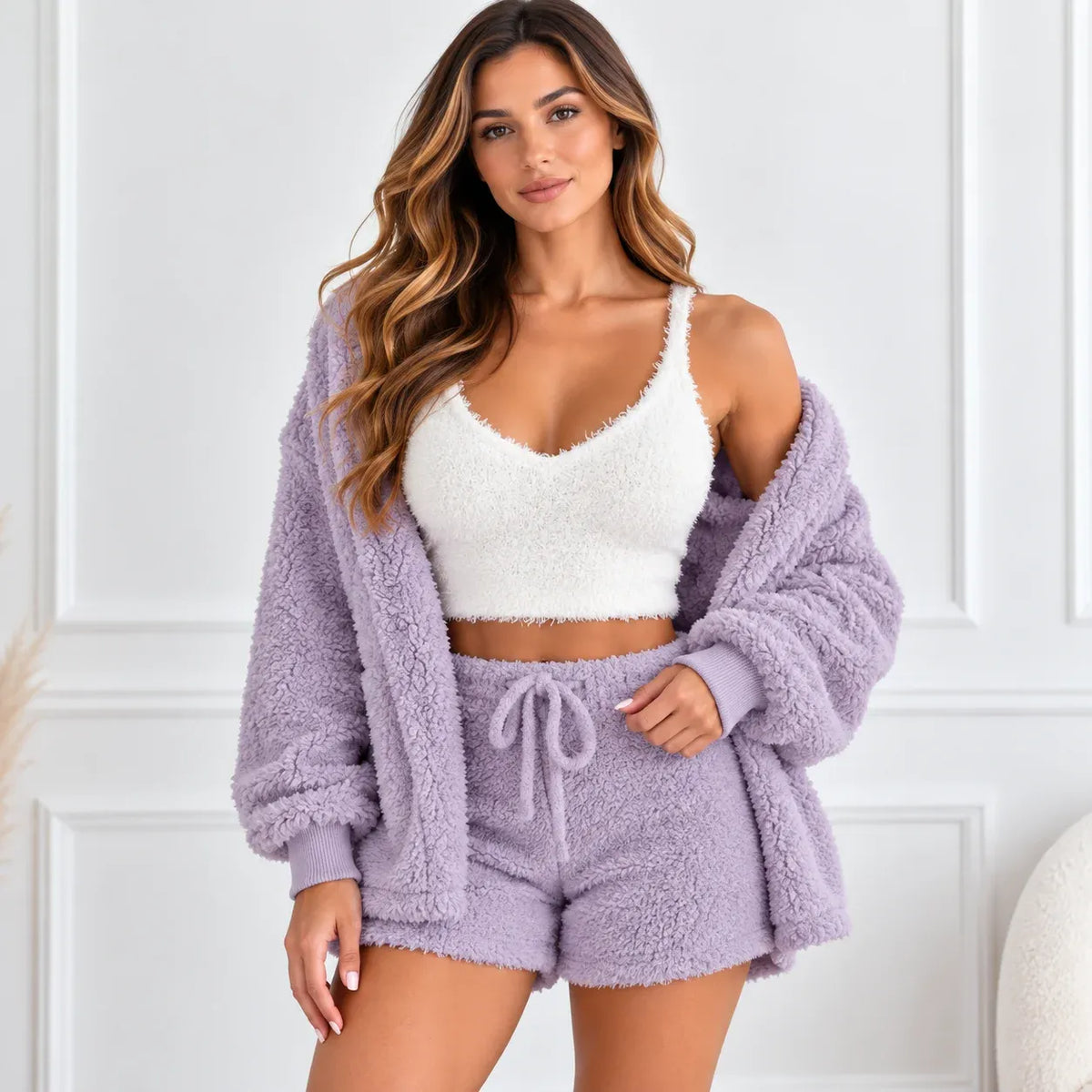 A woman models a cozy two-piece loungewear set featuring a lavender fuzzy sherpa cardigan and matching shorts over a white fuzzy camisole, set against a white paneled wall.