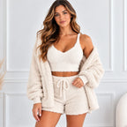 A woman modeling a cozy cream-colored three-piece loungewear set consisting of a cropped tank top, matching shorts with a drawstring, and a plush open-front cardigan, set against a light paneled wall.